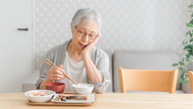 頬に手を当てて箸を止めている高齢女性